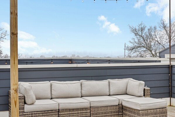 A sectional sofa with a wicker frame and cushions, positioned on a rooftop terrace with a view of the city skyline.