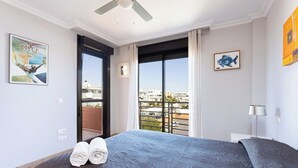 A hotel room with a bed, a ceiling fan, a large window with a view of the city, and artwork on the walls.