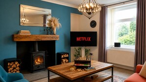 Een gezellige woonkamer met een open haard, een salontafel, een bank, een televisie waarop 'NETFLIX' wordt weergegeven, en een groot raam met gordijnen.