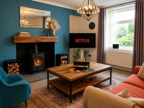 Ett mysigt vardagsrum med en öppen spis, ett soffbord, en soffa, en tv-skärm som visar ”NETFLIX” och ett stort fönster med gardiner.