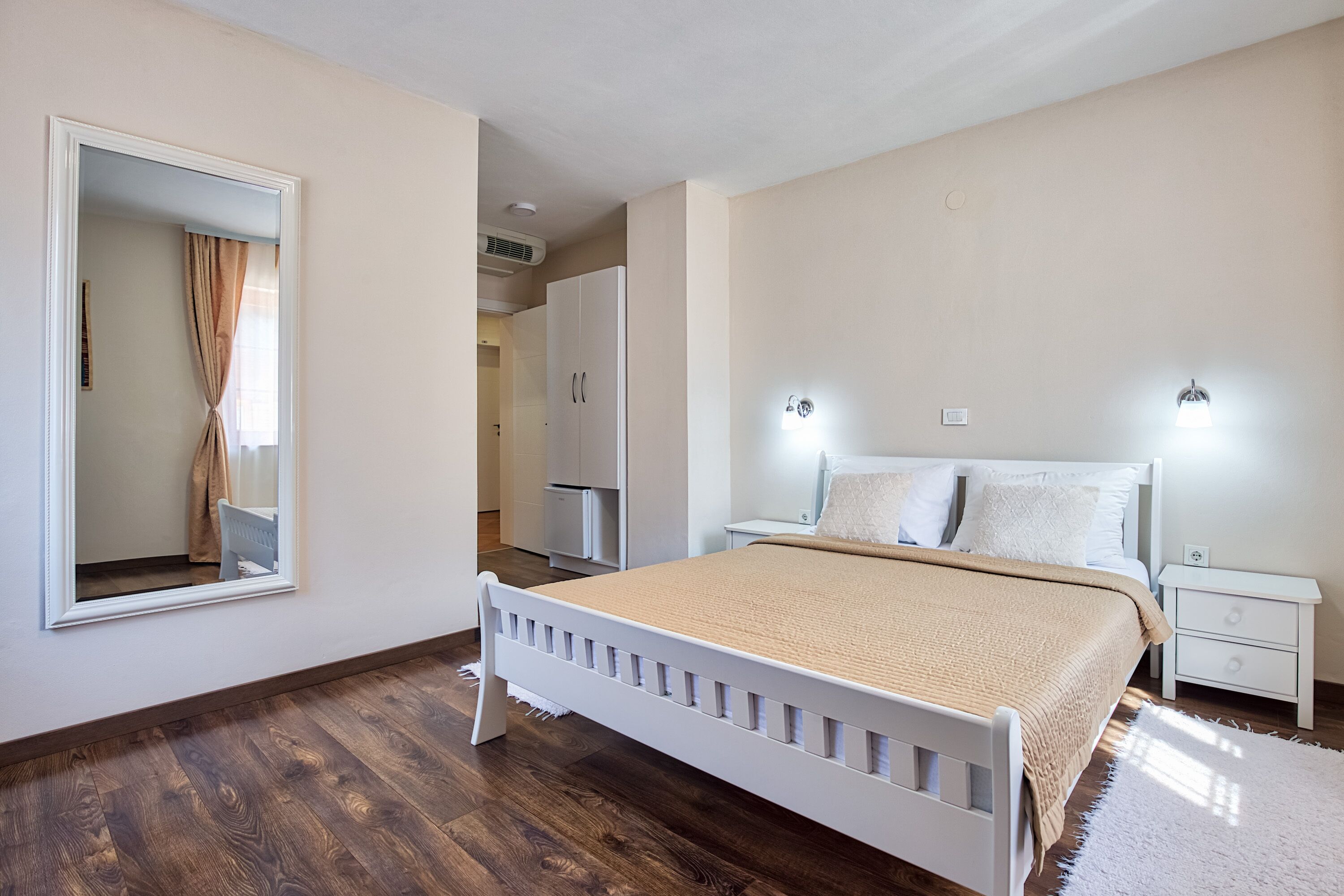 A bedroom with a wooden floor, a bed with a beige blanket, a nightstand, a mirror, and a wardrobe.