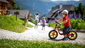 A child riding a tricycle on a gravel path with houses and mountains in the background. - Berg- & Erlebnis-Resort MarktlAlm - Chalets & Lodges (Stadl-Predlitz)