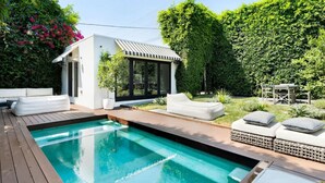 Una piscina con cubierta de madera, un pequeño edificio blanco con toldo a rayas y setos verdes exuberantes.