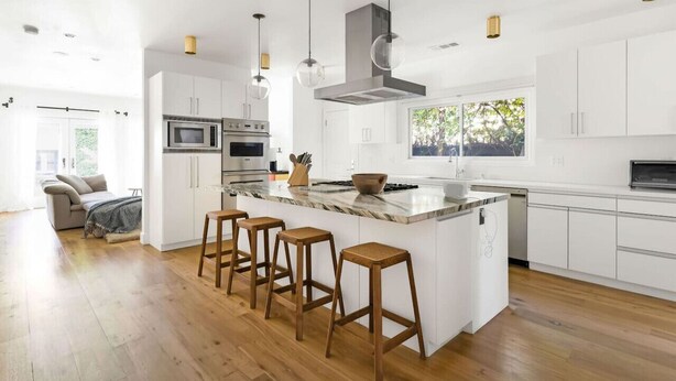 Una cocina moderna con armarios blancos, una isla central con taburetes, electrodomésticos de acero inoxidable e iluminación colgante.