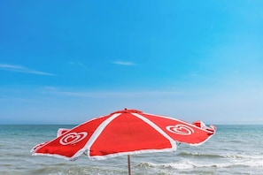 Un parasol de plage aux motifs rouges et blancs, placé sur le rivage d’une plage, avec un ciel bleu dégagé.