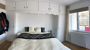 Een slaapkamer met een bed, een witte kast en een raam met uitzicht op gebouwen.