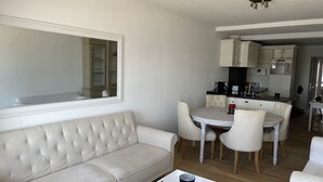 Een moderne woonkamer met een witte bank, een eettafel met stoelen, een keukenhoek en een grote spiegel aan de muur.