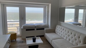 Een kamer met een groot raam met uitzicht op het strand, een witte bank, een kleine tafel en een gele stoel.