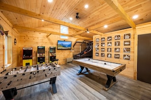 Sala com piso de madeira, mesa de pebolim, dois jogos de fliperama, mesa de bilhar e uma parede com ícones de atividades.