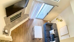 Een moderne woonkamer met een grijze bank, een witte salontafel en een aan de muur bevestigde televisie. Er is een trap die naar een hoger gelegen verdieping leidt, en een raam met gordijnen.