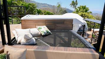 A balcony with a glass enclosure, wooden flooring, and a bed with white bedding and a rolled towel.