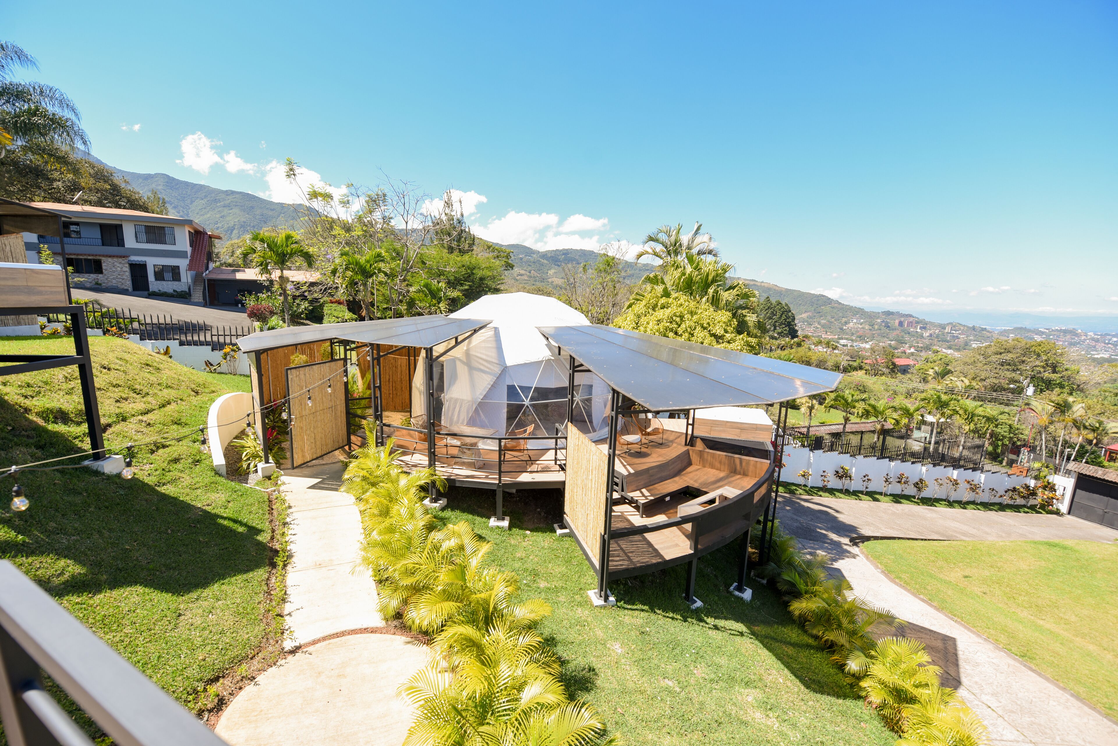 A modern outdoor seating area with a transparent roof, surrounded by lush greenery and a view of distant mountains.