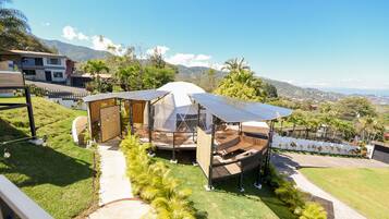 A modern outdoor seating area with a transparent roof, surrounded by lush greenery and a view of distant mountains.