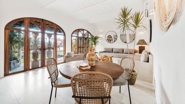 Amplia sala de estar con una mesa redonda de madera, sillas de mimbre, un sofá y ventanas grandes con vistas a la vegetación.
