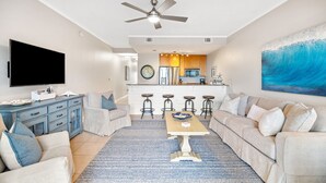 Un salon moderne avec un canapé, une table basse et un téléviseur. On aperçoit une cuisine à l’arrière-plan, avec un tabouret de bar et un tableau représentant une vague.