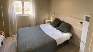A bedroom with a bed, a nightstand, a bedside table, a window with curtains, and a wooden ceiling. - The family cabin (Champdieu)