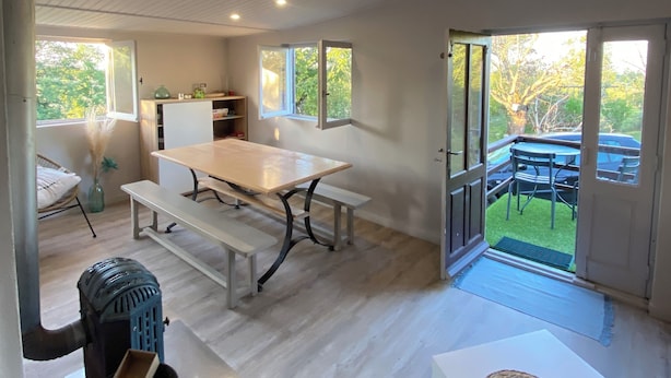 A spacious room with a wooden floor, a large table with benches, a small table with a woven placemat, and a view of a garden through a glass door. - The family cabin (Champdieu)