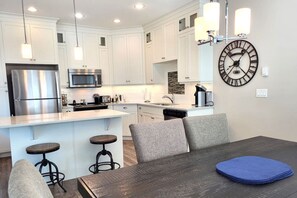 A modern kitchen with white cabinets, stainless steel appliances, a central island with stools, and a large wall clock.