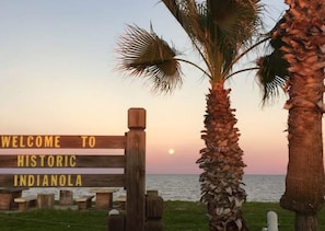 Uma placa de madeira com a inscrição 'WELCOME TO HISTORIC INDIANOLA', com vista para um corpo de água e palmeiras ao primeiro plano.