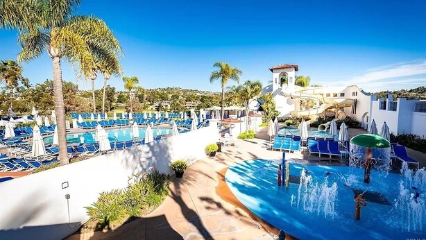 Een resort met een groot zwembad, palmbomen en een wit gebouw met een rood dak.