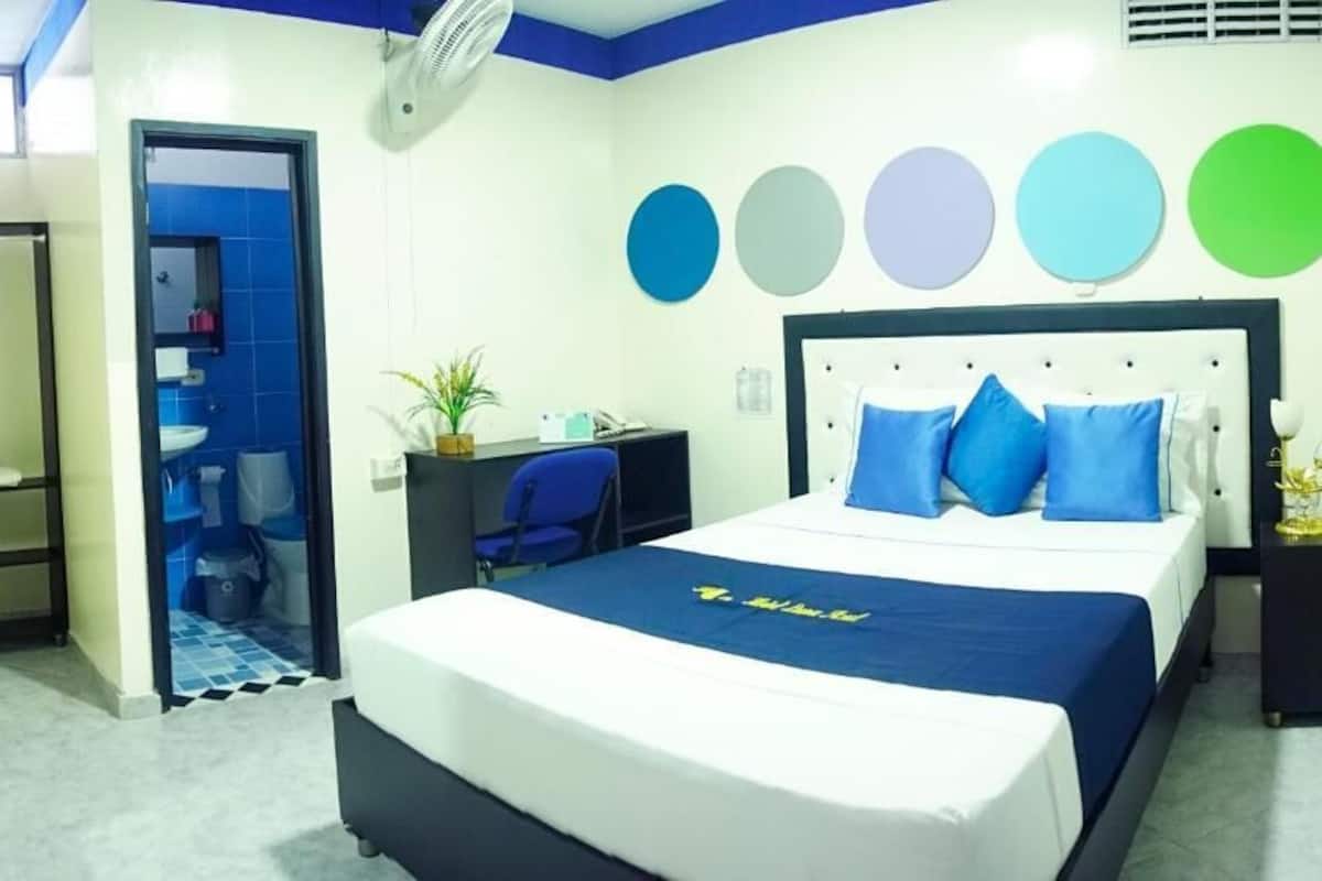 A hotel room with a bed, a desk, a chair, and a small table. There is a bathroom visible through an open door.