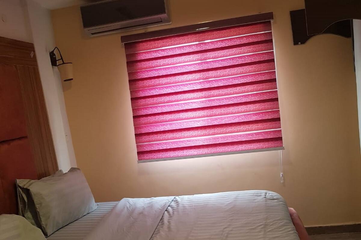 A bedroom with a bed, pillows, a window with pink blinds, an air conditioning unit, and a wooden wardrobe.