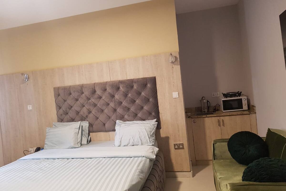 A hotel room with a large bed, a wooden headboard, a small kitchenette with a microwave, and a green armchair.