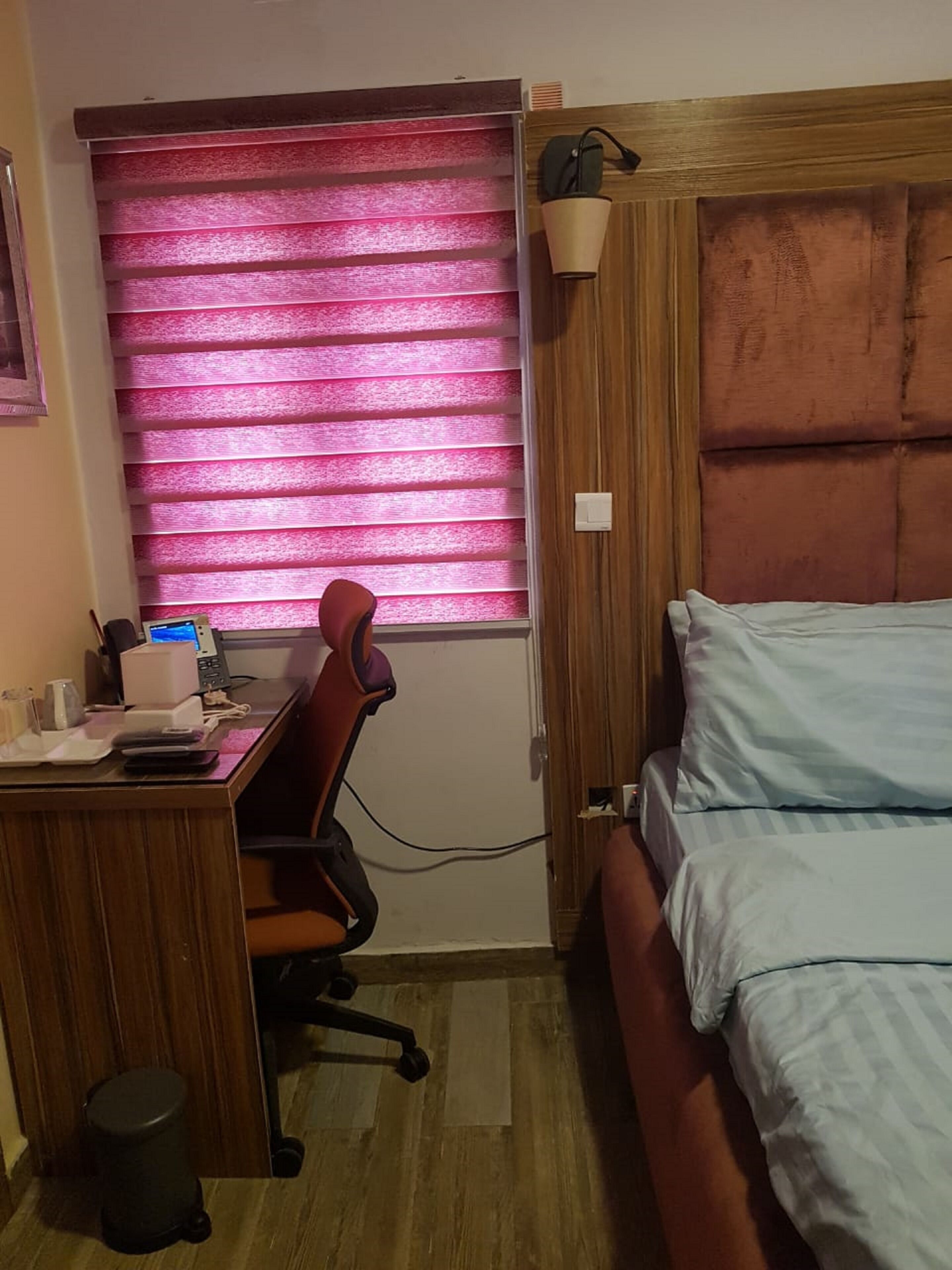 A bedroom with a bed, a desk with a chair, a window with pink blinds, and a wooden headboard.