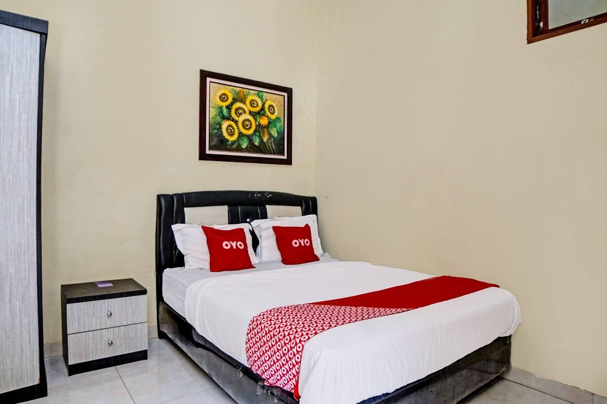 A neatly made bed with white bedding and red accents, a small bedside table with a lamp, and a framed painting of sunflowers on the wall.