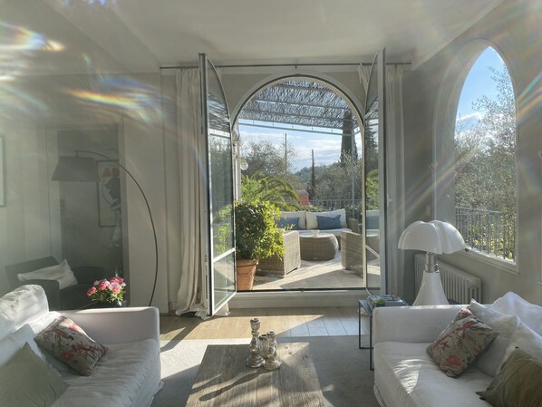 A sunlit room with a view of a balcony, furnished with a sofa, a table, and a lamp.