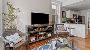 A living room with a television, wicker chairs, a bookshelf, and a kitchen area with white cabinets and appliances. - Cozy Convenient downtown Condo (Wilmington)