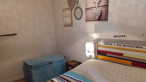 A bedroom with a bed, a bedside table, a lamp, a wall decoration, and a blue suitcase. - Pretty little house, in a quiet private courtyard 5 minutes from the sea (Luc-sur-Mer)