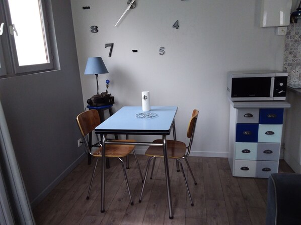 A small room with a table and chairs, a lamp, a microwave, and a cabinet with drawers. - Pretty little house, in a quiet private courtyard 5 minutes from the sea (Luc-sur-Mer)