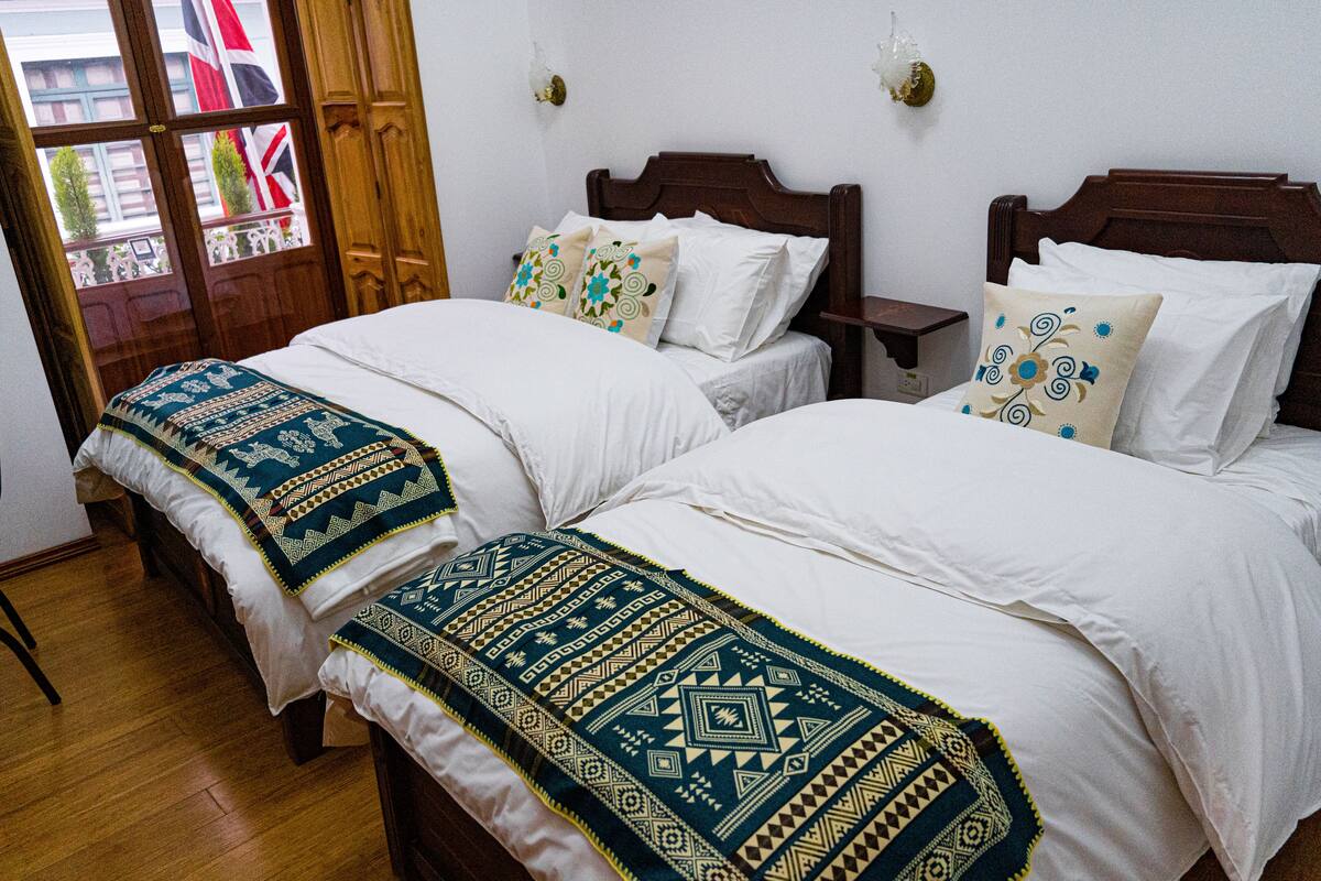 Habitación de hotel con dos camas, cada una con colcha y almohadas estampadas, cabeceros de madera y una mesita de noche con lámpara.