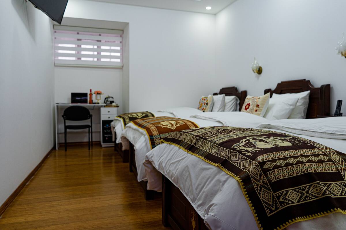 Habitación de hotel con tres camas individuales, un escritorio con silla, una mesita y una ventana con persianas.