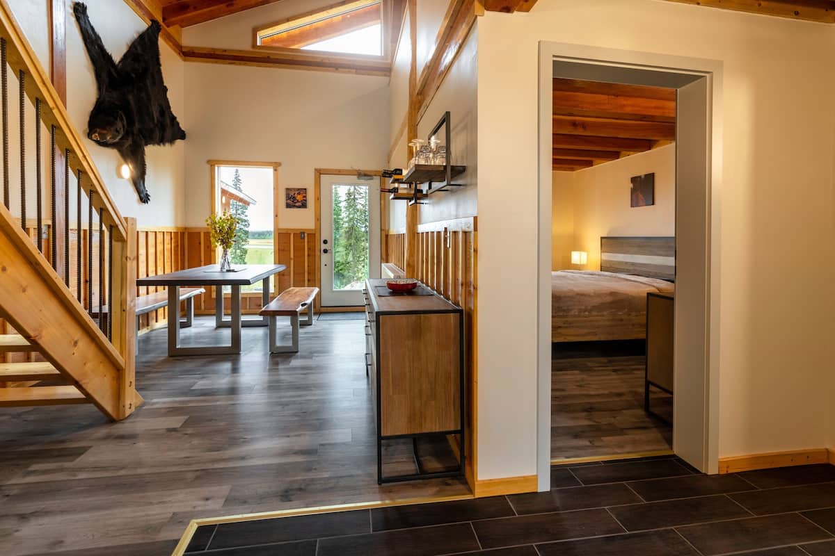 A spacious room with a wooden staircase, a dining area with a table and benches, a kitchen area with a countertop and sink, and a bed with a headboard.
