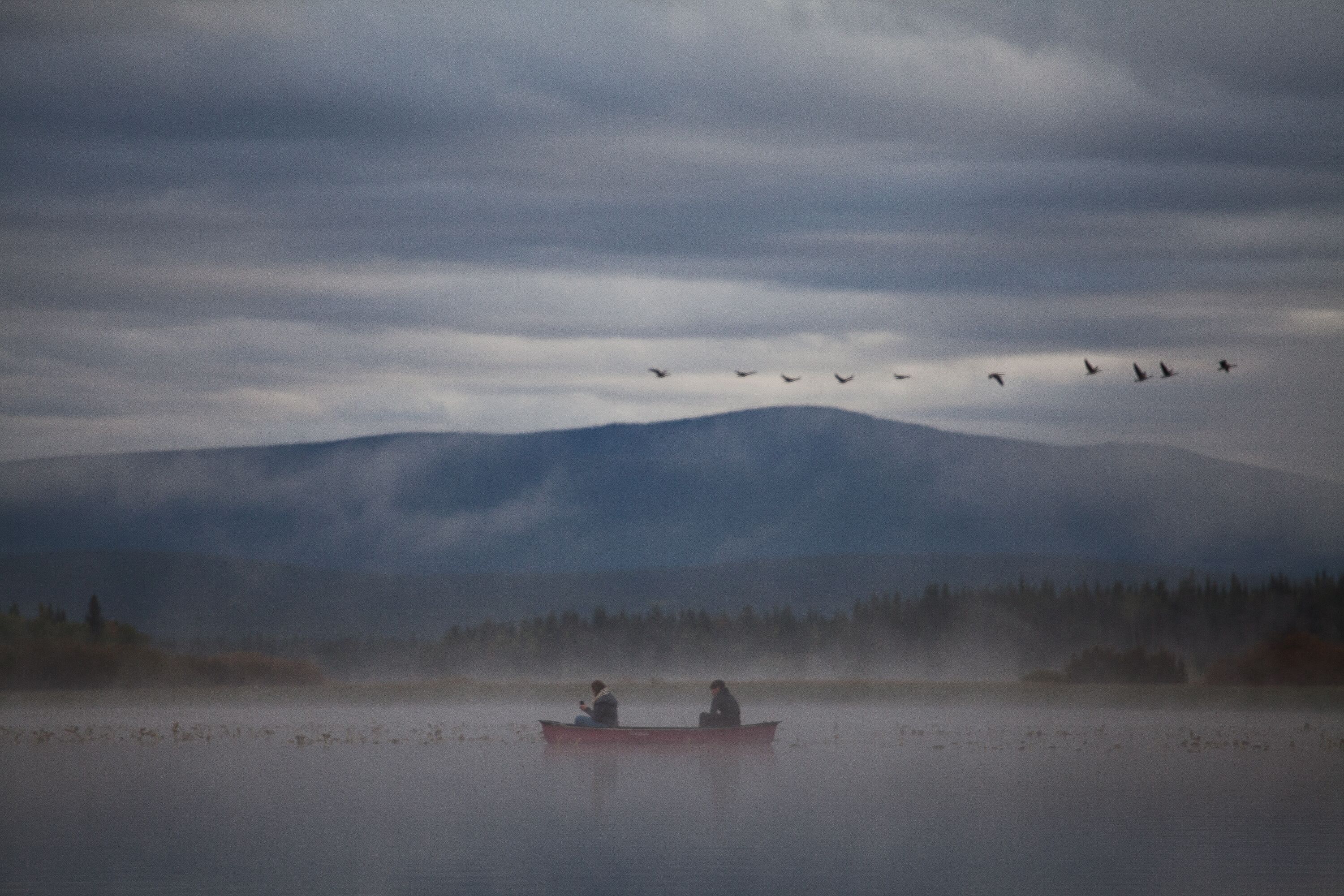 A misty lake with a boat carrying two people, surrounded by mountains and a flock of birds in the sky.