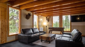 A cozy living room with wooden beams, a leather sofa, a coffee table, and a foosball table. - Red Cariboo Resort (Anahim Lake)