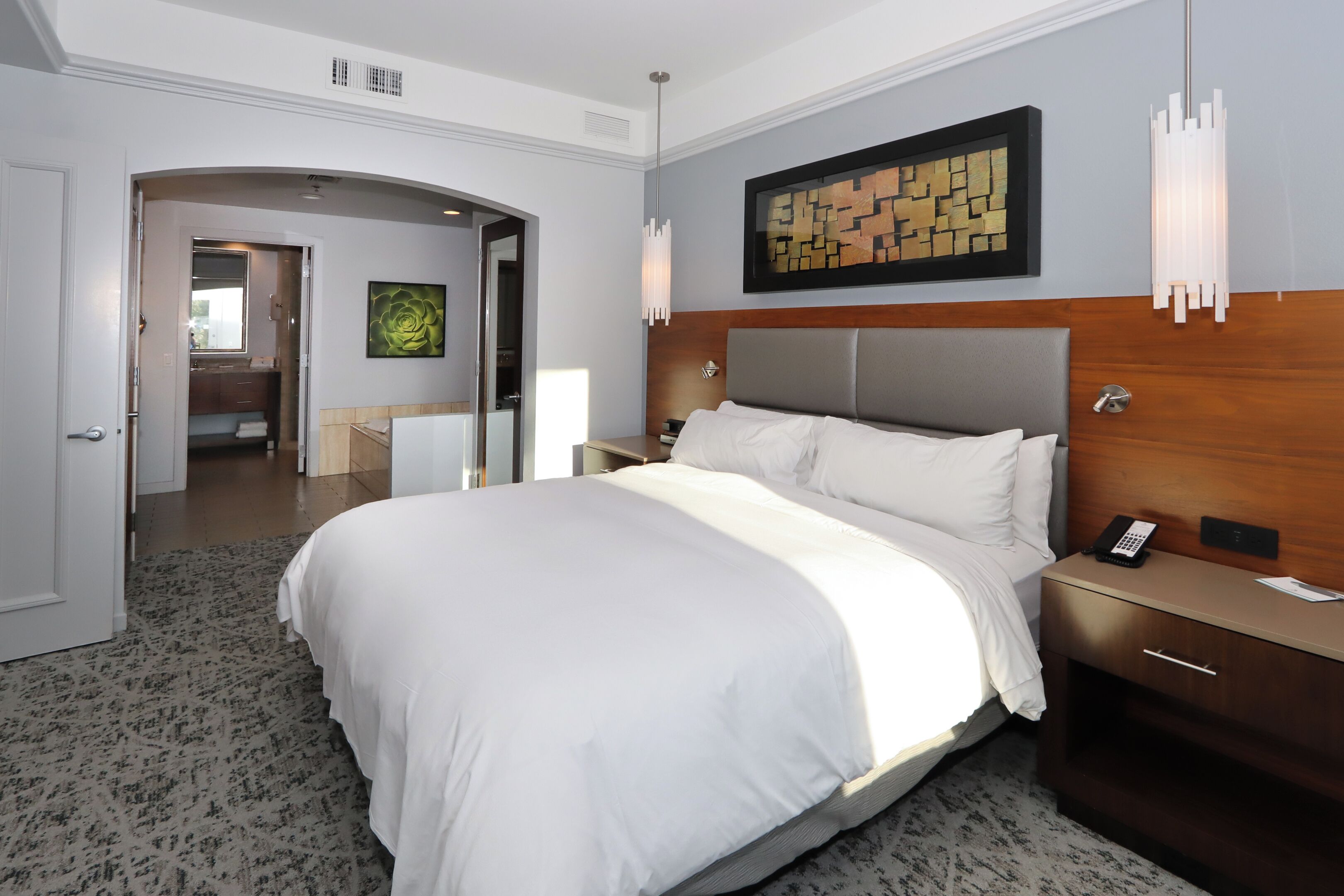 A hotel room with a large bed, a wooden headboard, a nightstand, a bedside table, a wall-mounted artwork, and a door leading to a bathroom.