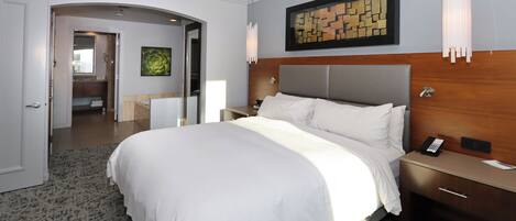 A hotel room with a large bed, a wooden headboard, a nightstand, a bedside table, a wall-mounted artwork, and a door leading to a bathroom.