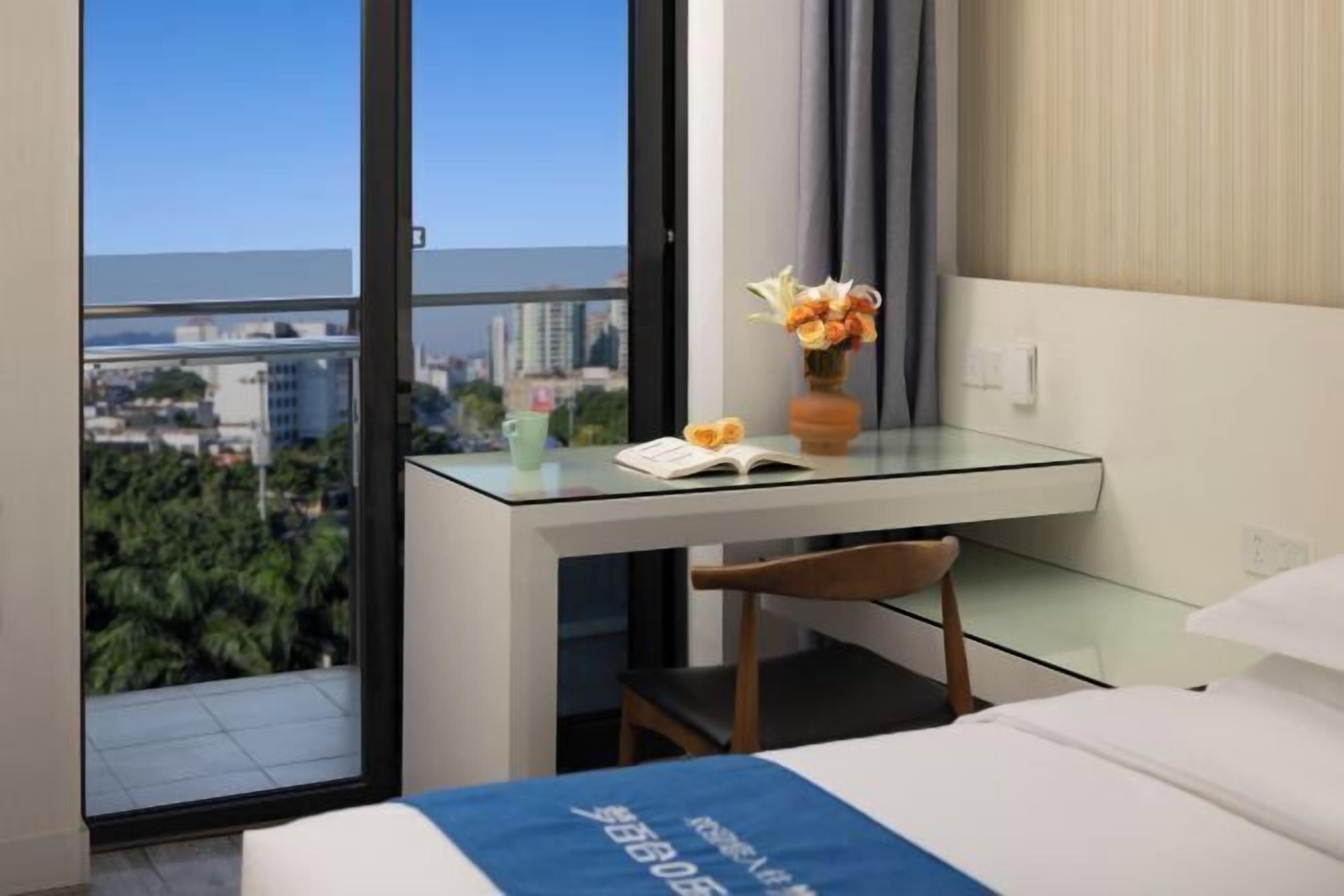 A hotel room with a glass desk, a chair, a bed, a vase with flowers, and a view of the city.