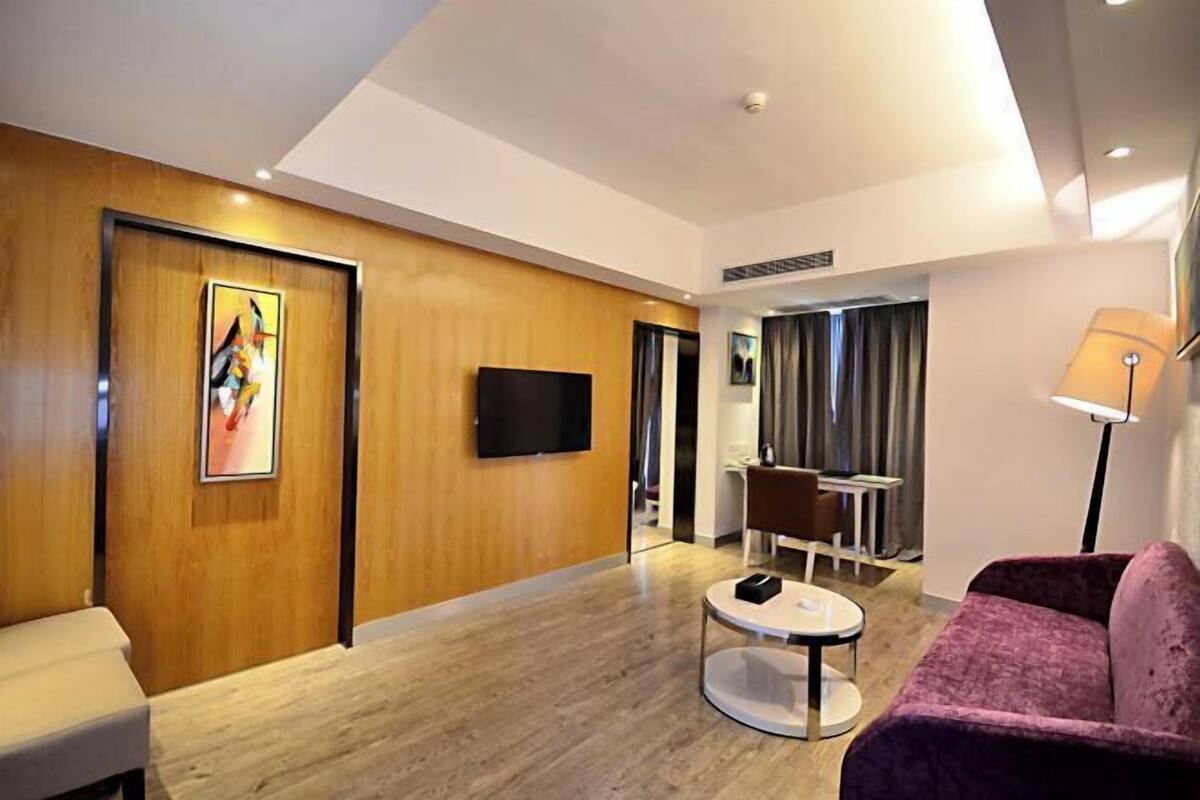 Une chambre d’hôtel moderne avec un mur d’accent en bois, un téléviseur à écran plat, une table basse ronde, un canapé violet et un coin repas avec une table et des chaises.