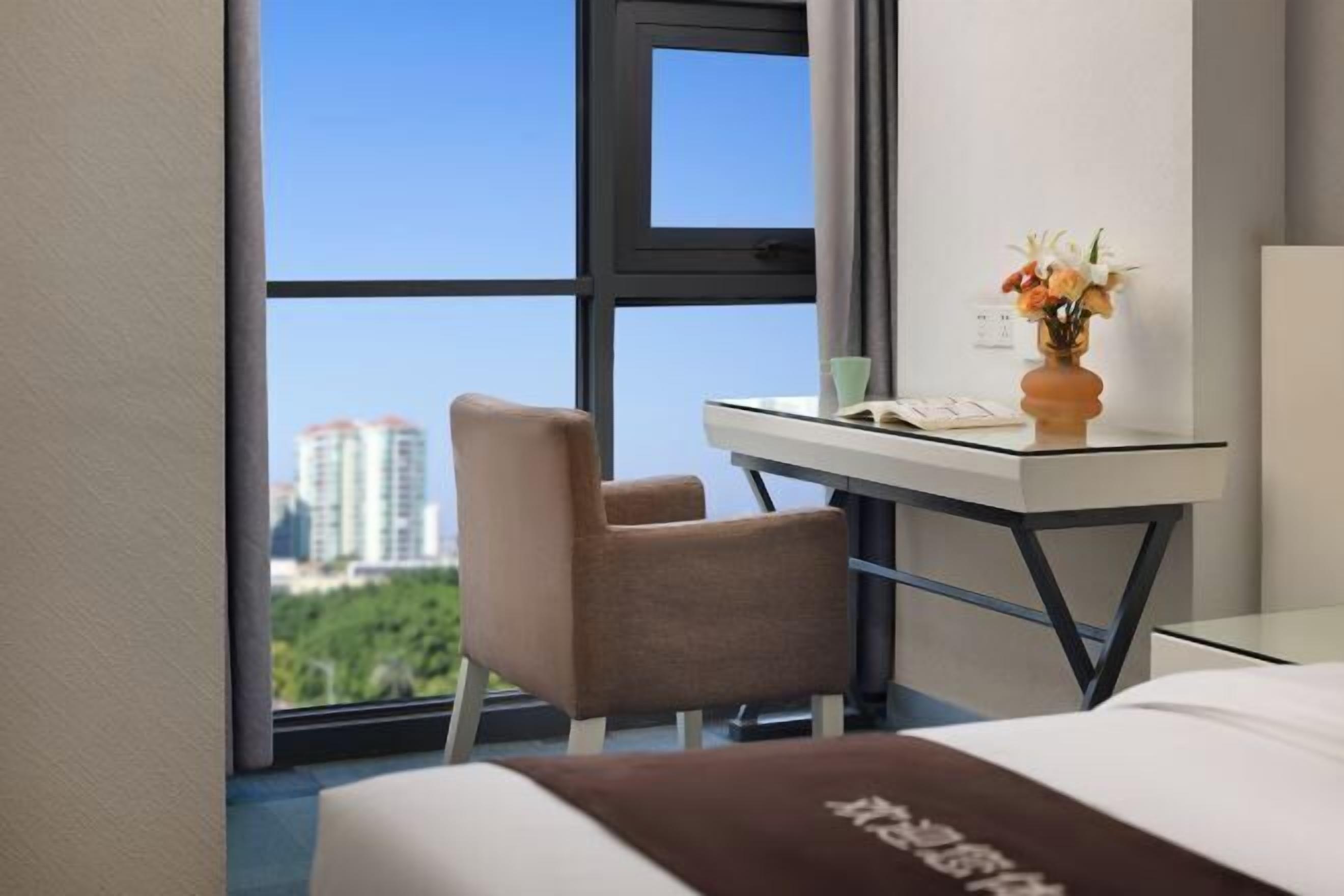 Una habitación de hotel con cama, escritorio con silla, florero con flores y ventana con vista a la ciudad.