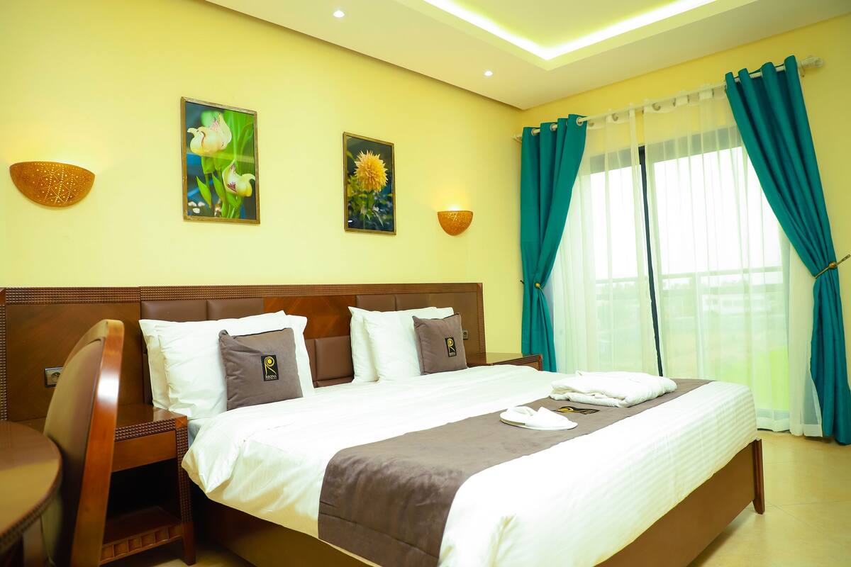 Une chambre d’hôtel avec un grand lit, une tête de lit en bois, un bureau avec une chaise et une fenêtre avec des rideaux.