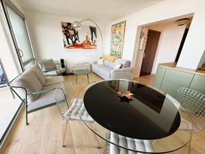 A modern living room with a glass table, a sofa, a chair, and a kitchen area.