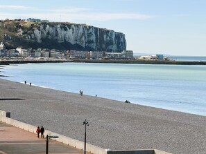 A pebble beach with a cliff in the background, a promenade, and a few people walking.