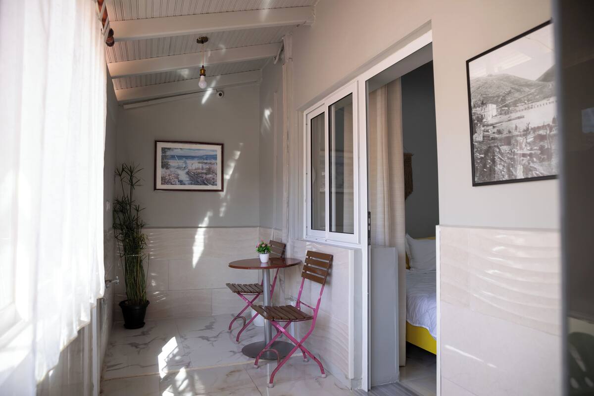 A small balcony with a table, chairs, a potted plant, and a bed in the background.