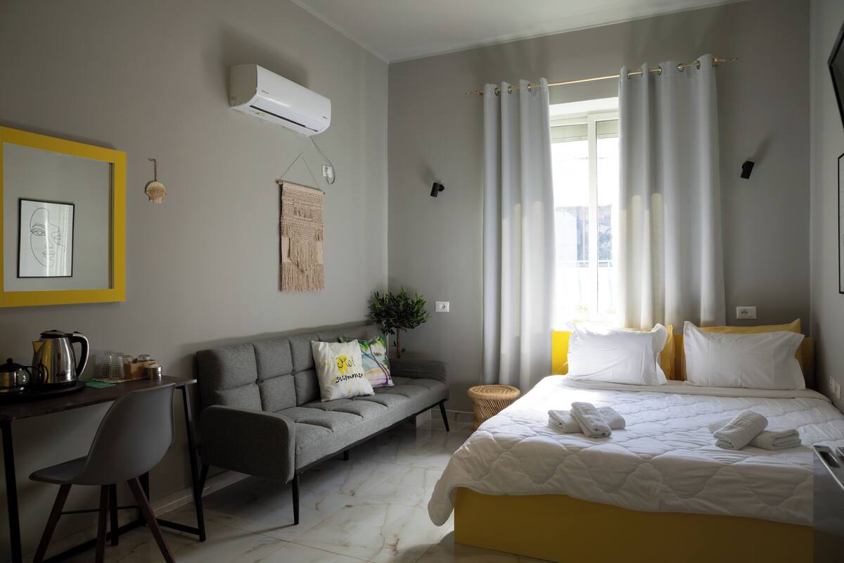 A modern hotel room with a bed, a sofa, a desk with a kettle, and a window with curtains.