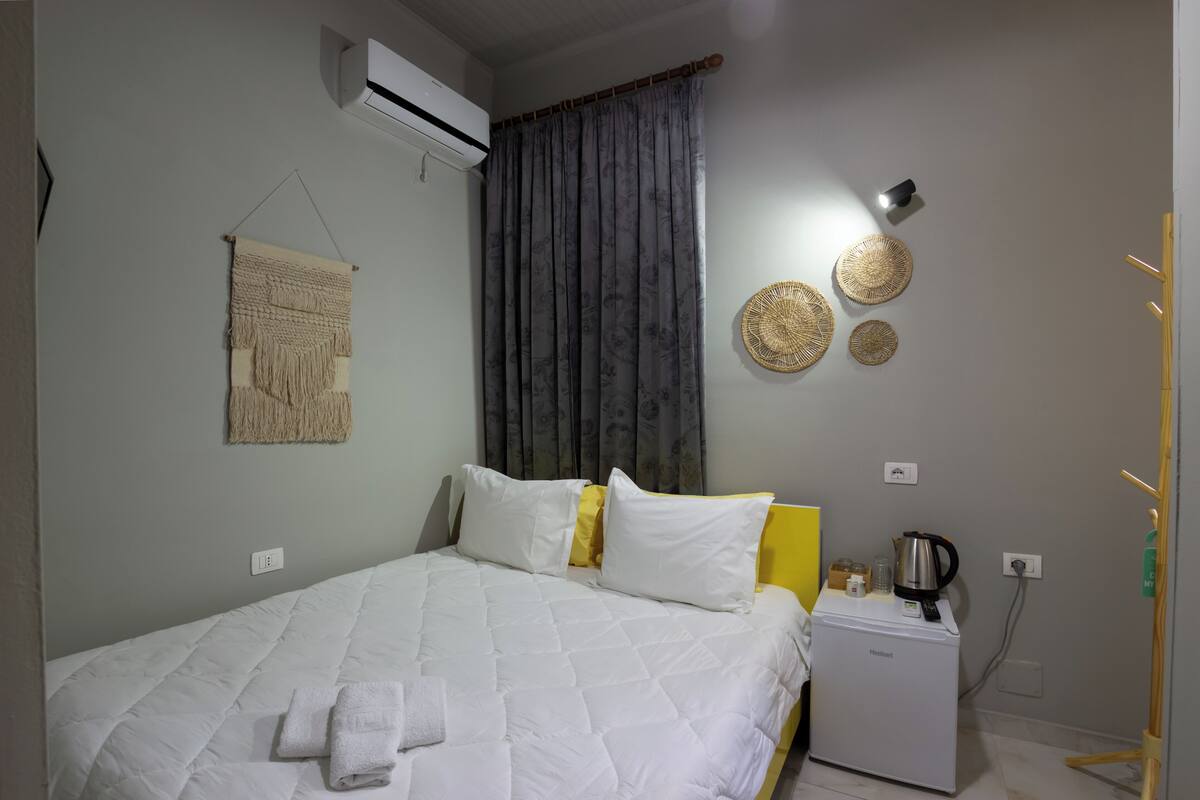 A neatly made bed with white and yellow bedding, a small white heater, and a wall-mounted air conditioner.
