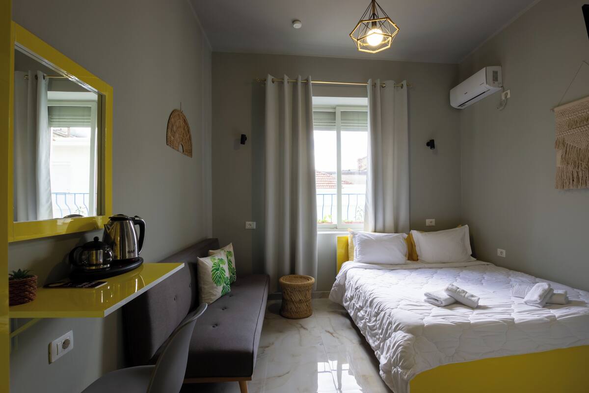 A hotel room with a bed, a bench, a small table with a kettle, a mirror, and a window with curtains.
